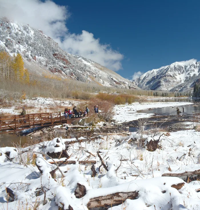 People on a footbridge over Maroon Creek near Aspen after the first snow, with snowy peaks and gold aspens behind